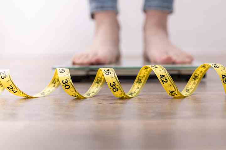 A close-up of a person’s feet standing on a scale, with a yellow measuring tape placed in the foreground, slightly out of focus. The measuring tape is coiled in a wave pattern across the wooden floor, emphasizing themes of weight measurement and body image.