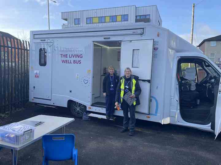 A white mobile health unit labelled ‘The Living Well Bus’ is parked outdoors with its side door open. Two staff members wearing uniforms and lanyards stand at the entrance of the vehicle. A small outdoor setup with a table, chairs, and storage box is positioned in front of the bus. Residential buildings and a metal fence are visible in the background.