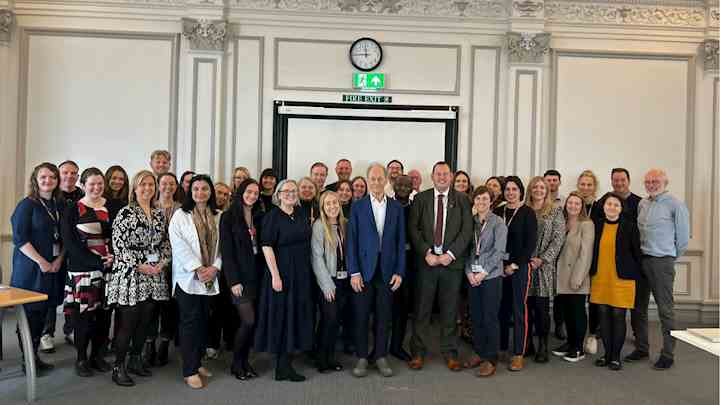 A group of Liverpool City Council staff members posing for a photo with Professor Sir Michael Marmot, who stands in the center wearing a blue suit jacket and white shirt. The group is gathered in a bright, formal room with ornate detailing on the walls, a clock, and an exit sign above a screen in the background