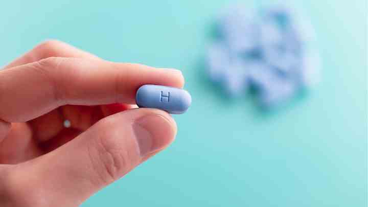 A close-up image of a hand holding a single blue pill with the letter "H" engraved on it, set against a soft teal background. In the blurred background, additional blue pills are visible.